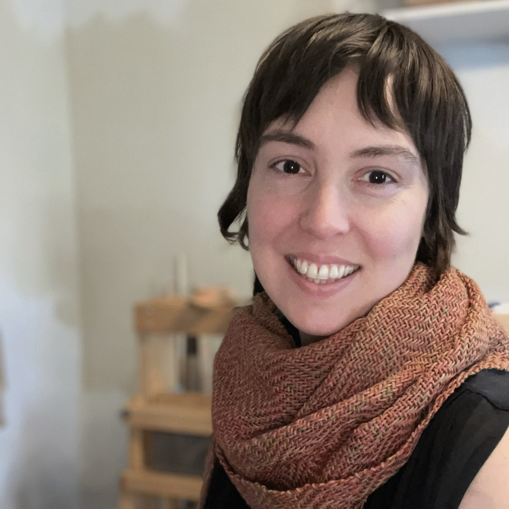 This is a photograph of Bea Huff Hunter, a smiling white woman in her forties with short brown hair. She is wearing an orange twill handwoven scarf and seated in front of a floor loom.